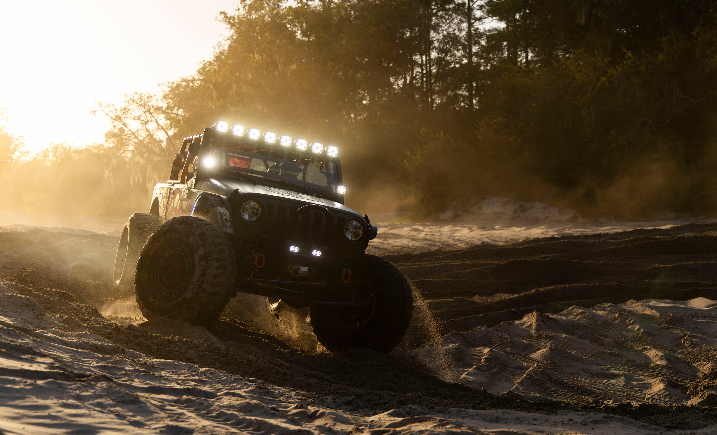 Jeep driving on a dirt road with lights on, surrounded by trees.