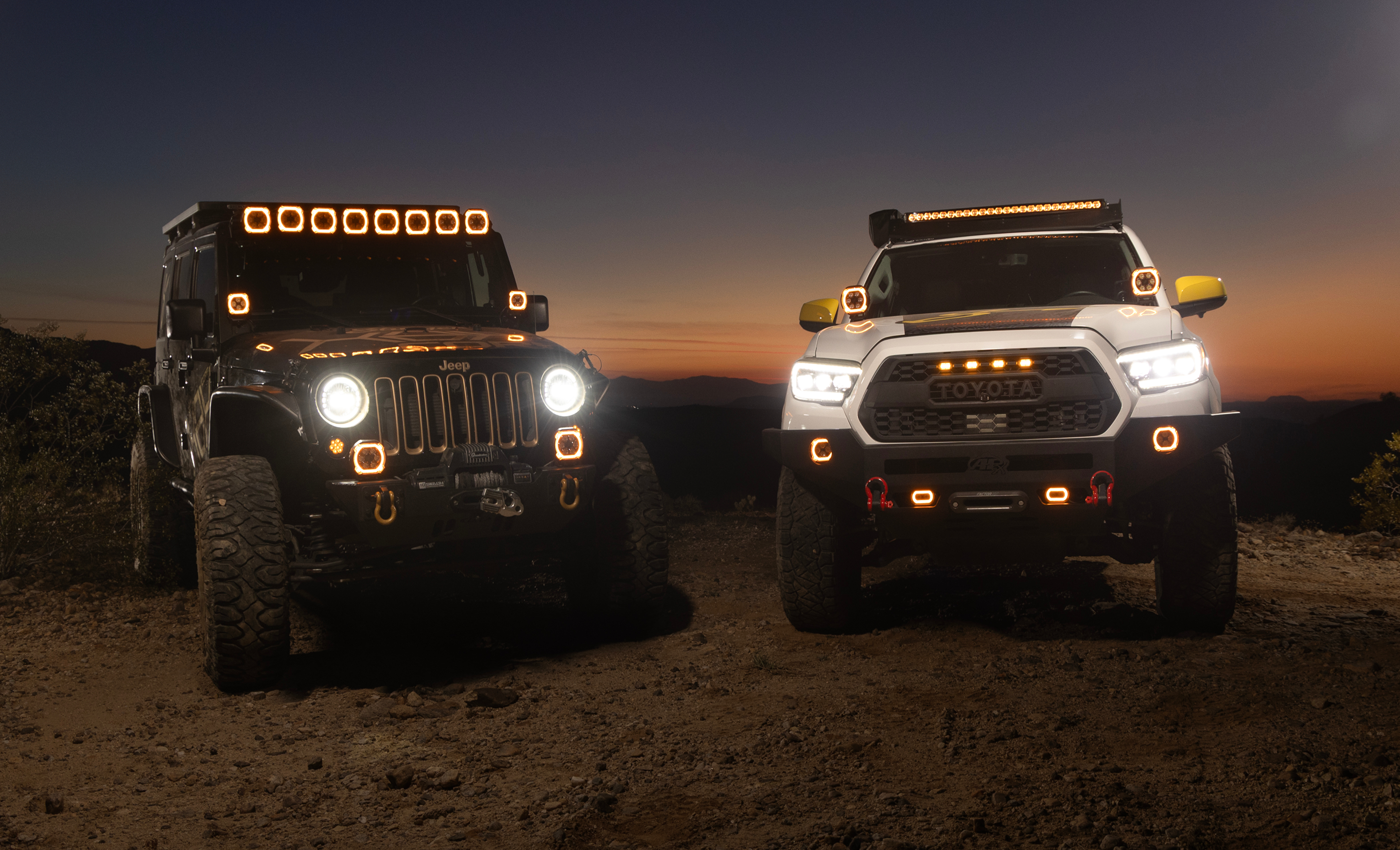 Two off-road vehicles, a Jeep and a Toyota Tacoma, are parked side by side on a dirt road at sunset.