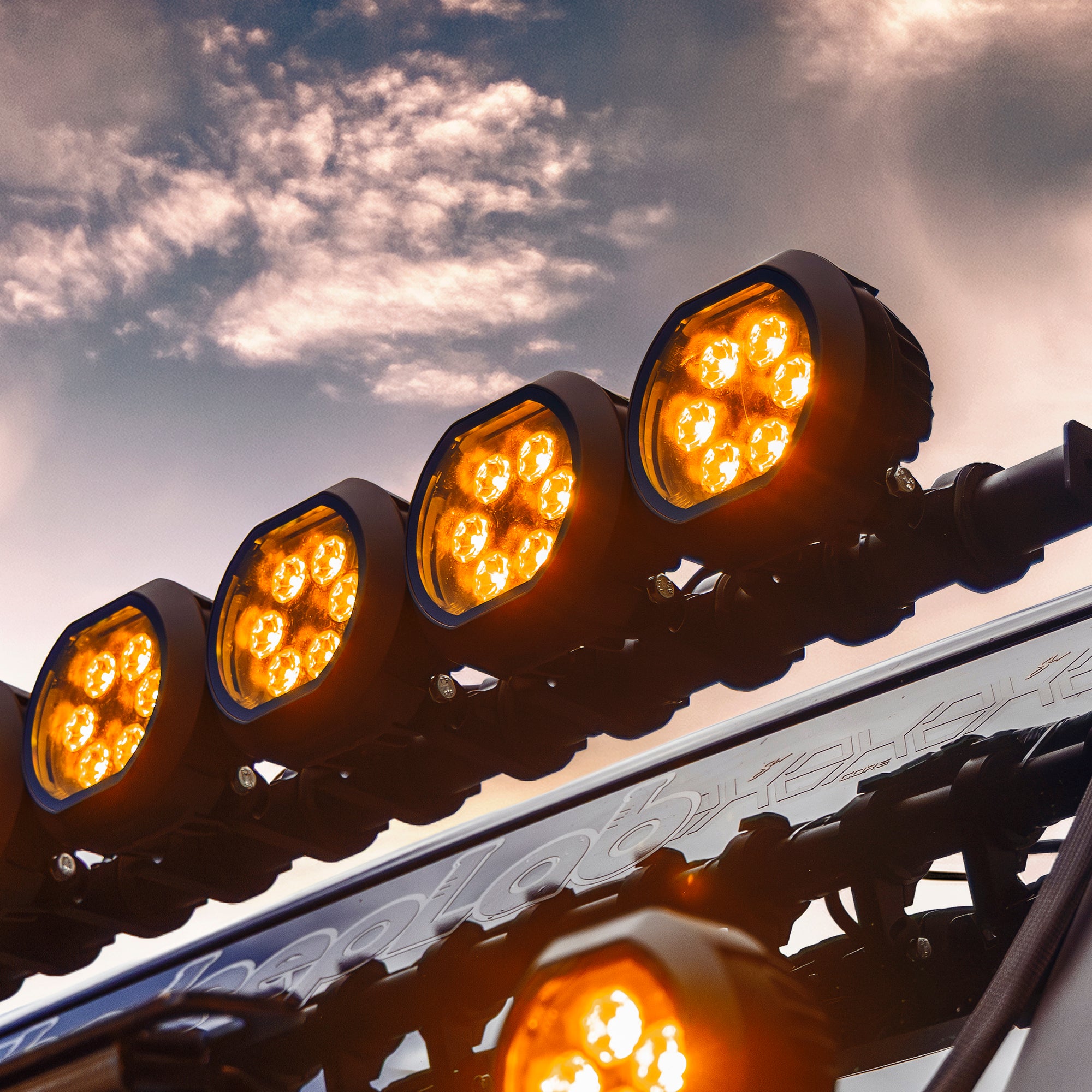 Row of four circular LED off road lights mounted on a vehicle roof rack against a cloudy sky