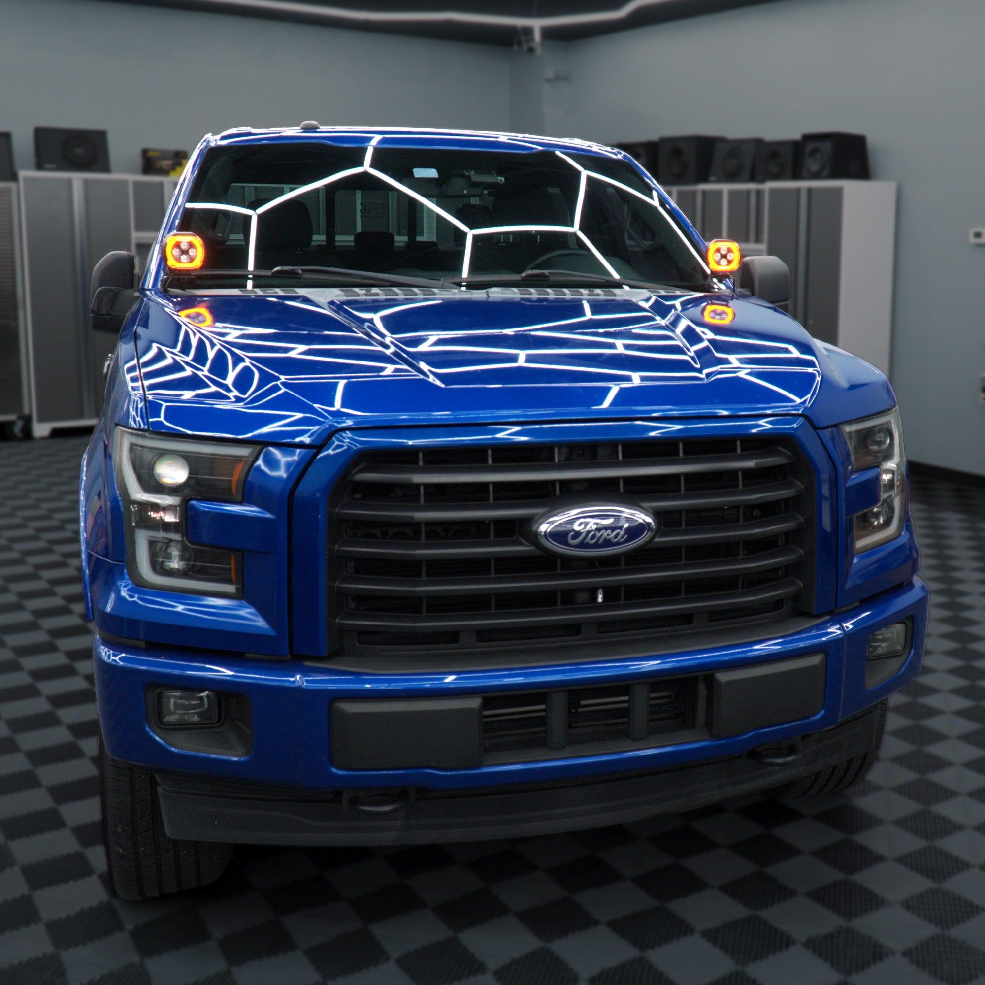 A blue Ford pickup truck photographed head-on inside a garage or studio. The windshield has a white geometric line pattern, and there are small orange-and-black flag decals near the outer sides of the hood.
