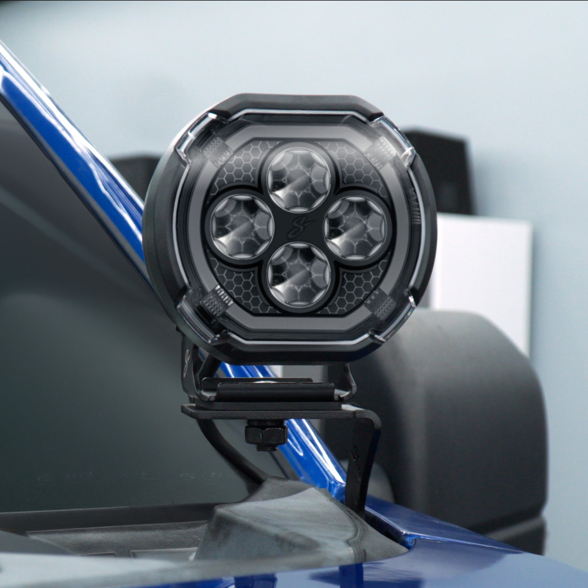 A black circular off-road LED light with four circular lenses, mounted on a blue vehicle frame.