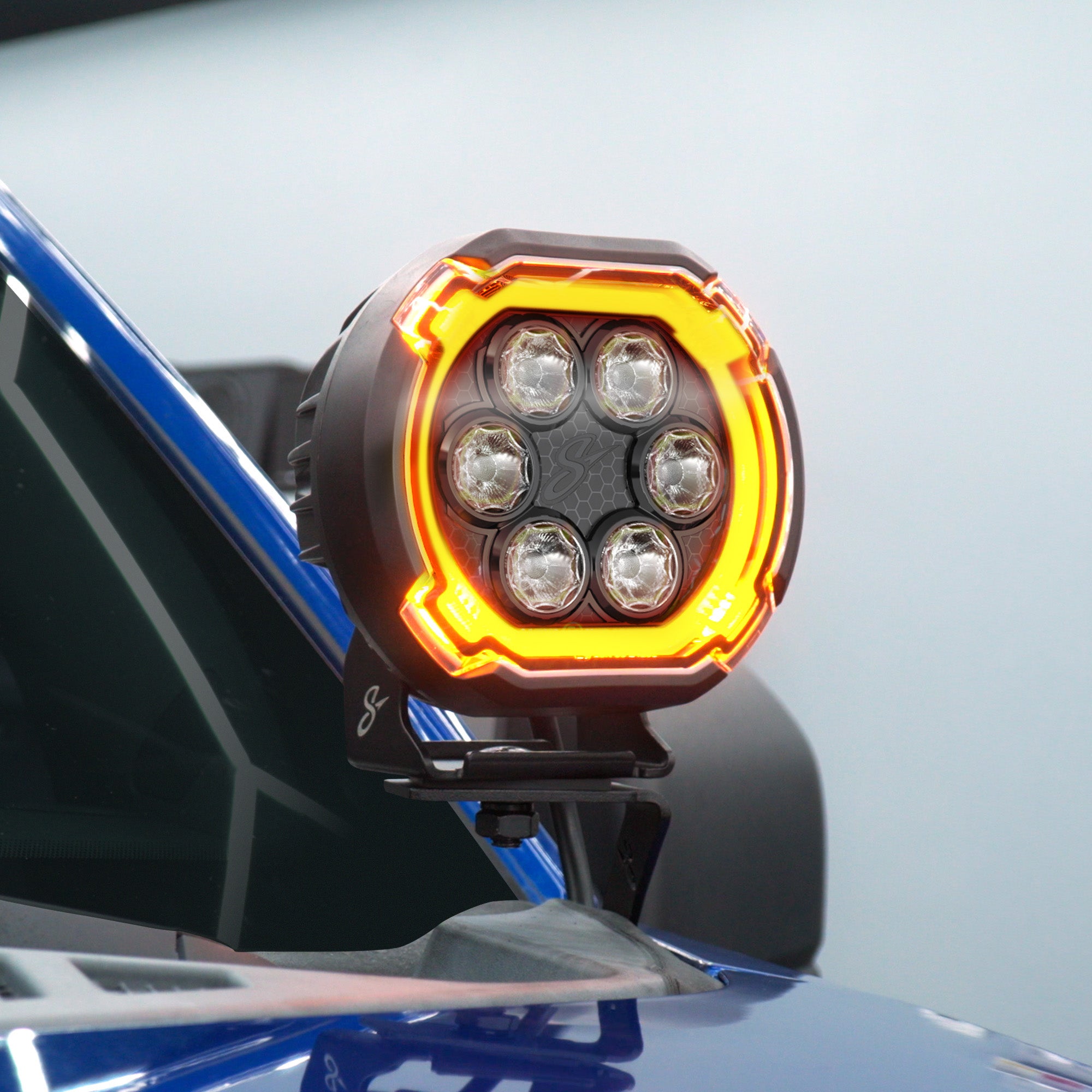 A close-up of a round automotive LED light mounted on a blue vehicle, with the light illuminated in orange and yellow.