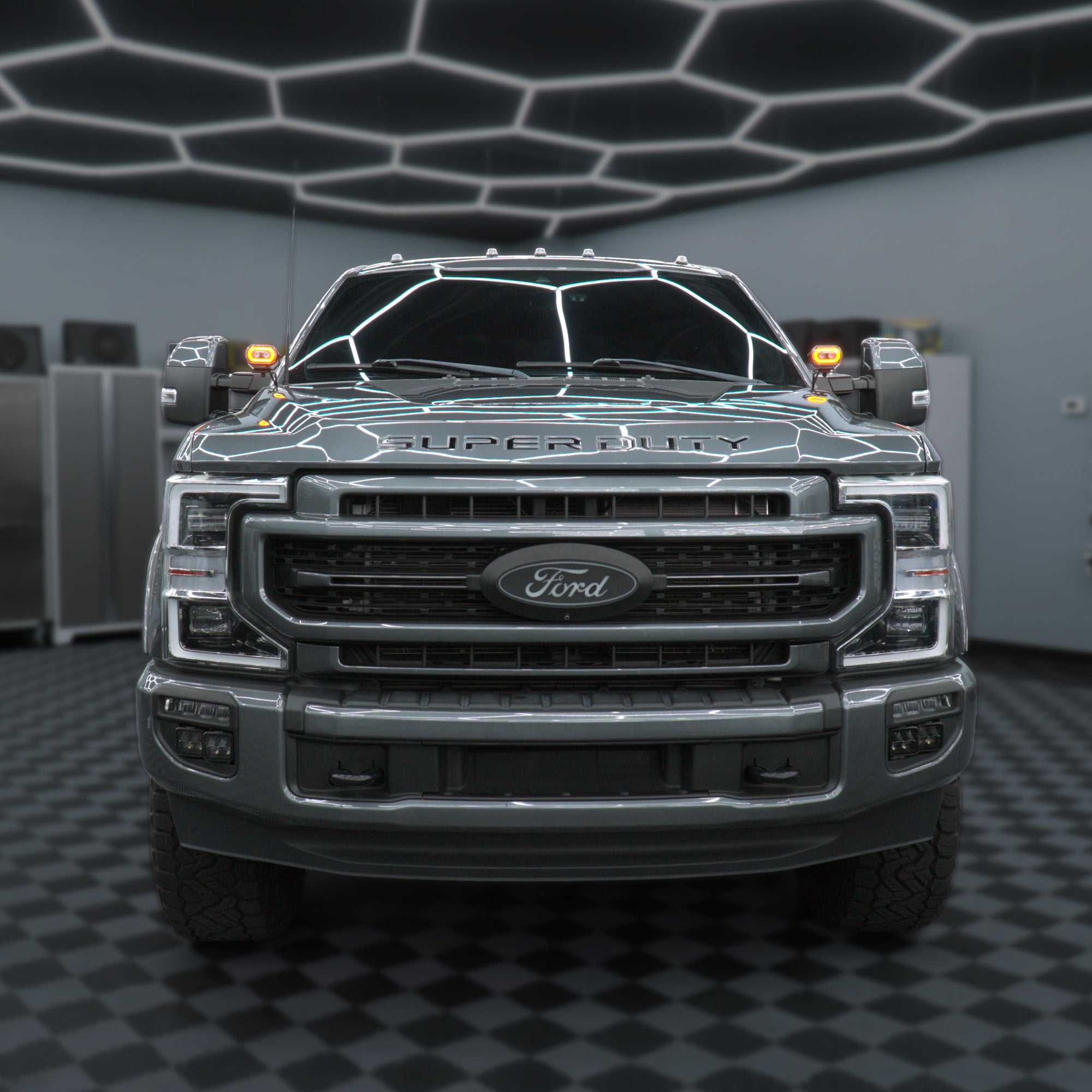 A front-facing view of a dark gray Ford Super Duty pickup truck parked inside a modern garage with a black-and-white checkered floor and geometric ceiling lights.