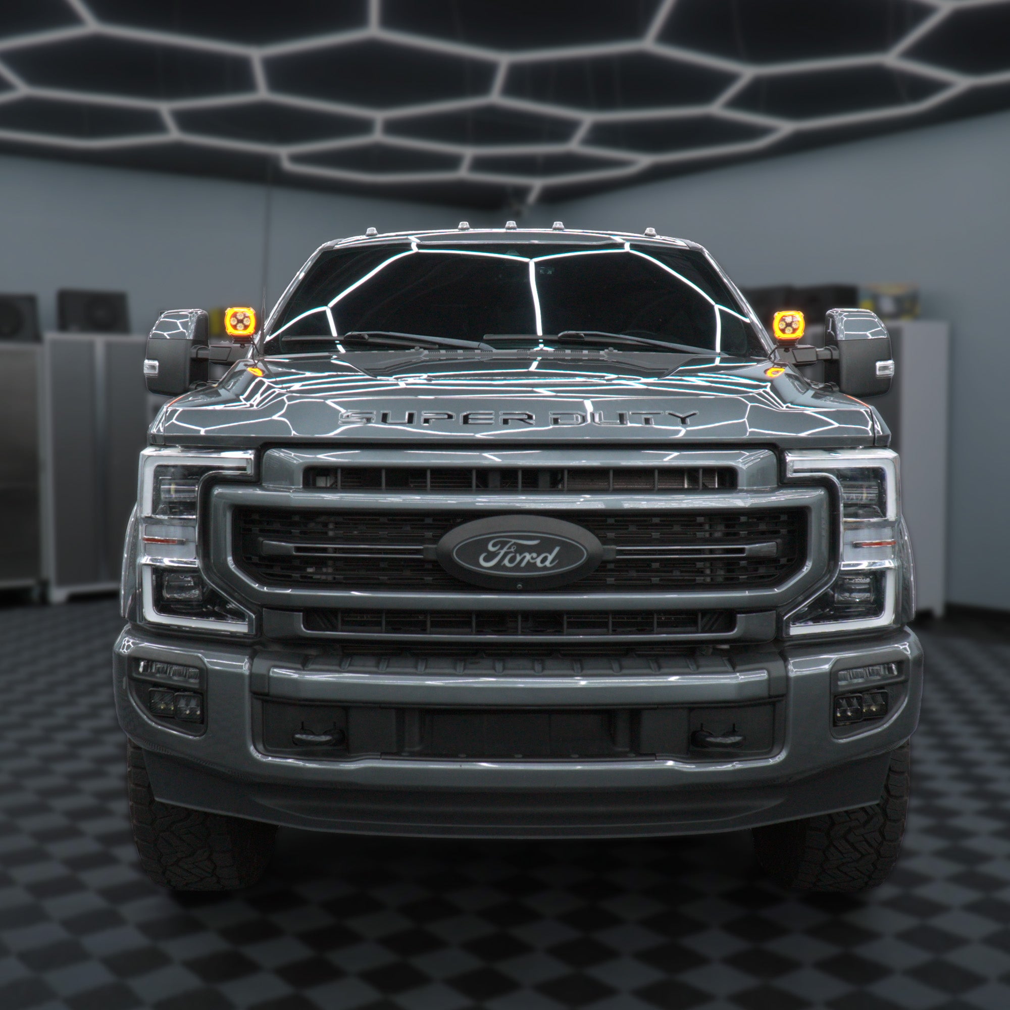 A front-facing view of a dark gray Ford Super Duty pickup truck parked in a showroom with a black-and-white checkered floor and geometric ceiling lights.