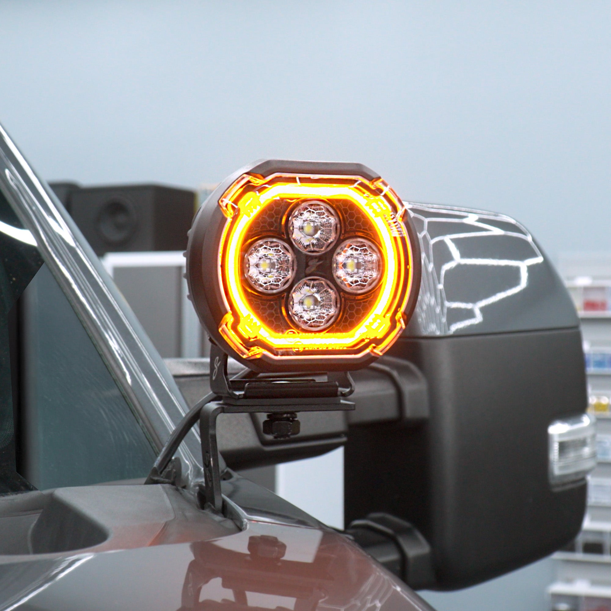 A close-up of a circular LED truck light mounted on a vehicle side mirror, with the amber light illuminated against a pale sky background.