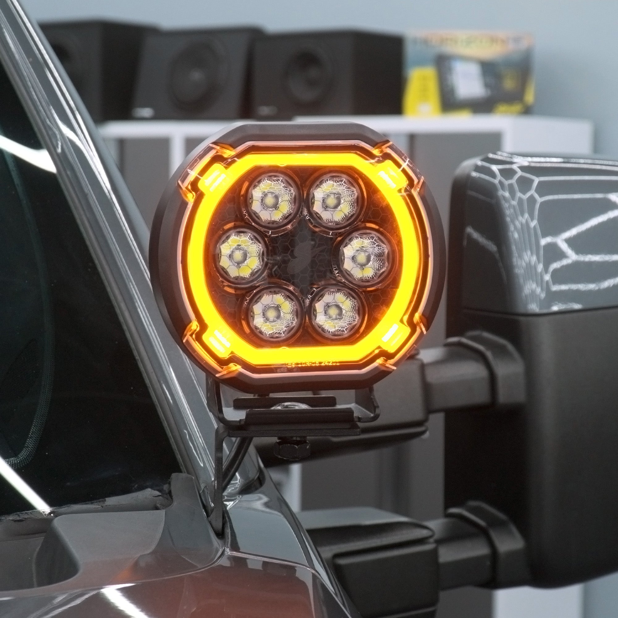 A close-up of a vehicle-mounted LED light bar with six illuminated LEDs and a bright amber light ring, attached to a dark vehicle side mirror.