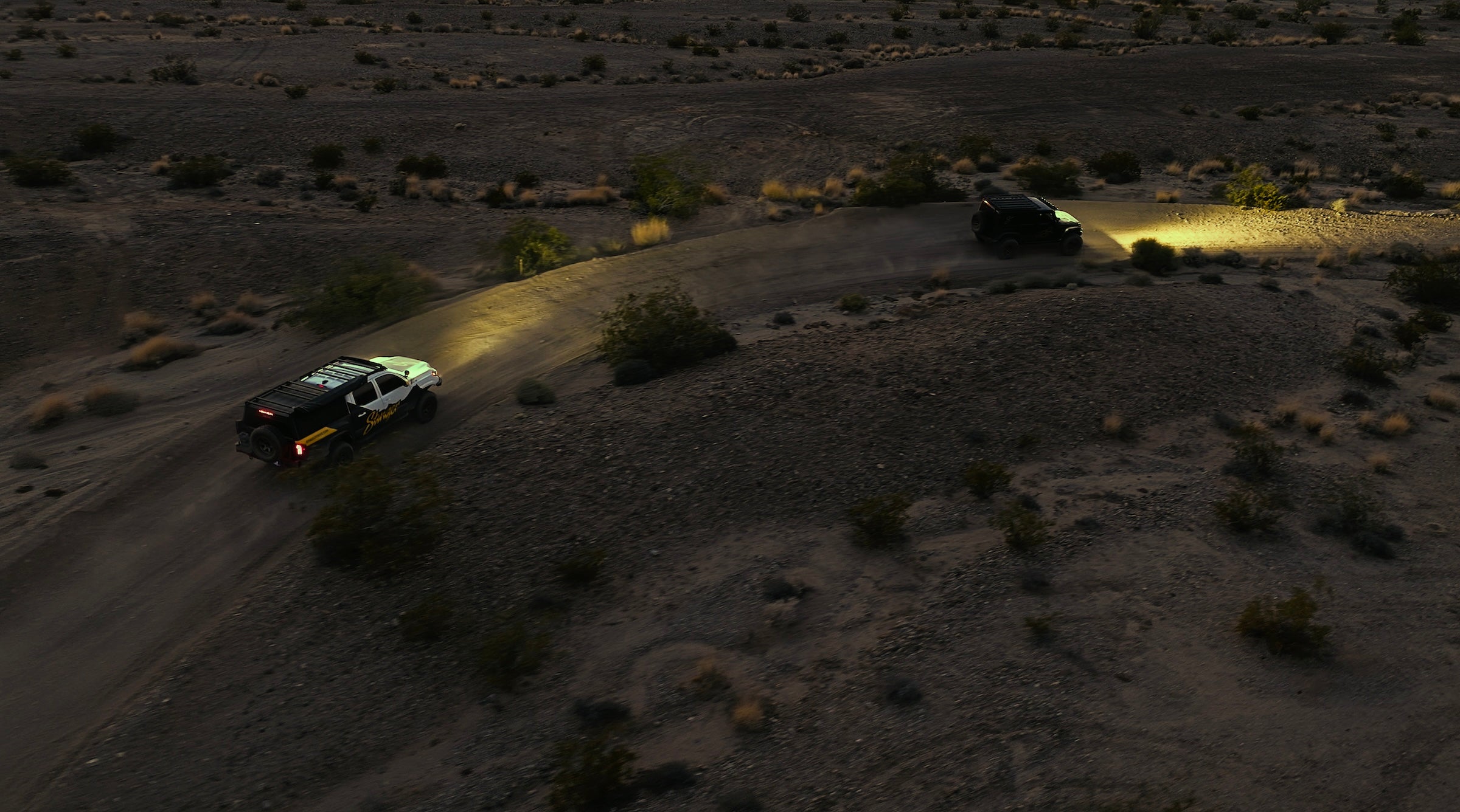 Car driving on a desert road with headlights on, surrounded by sparse vegetation.