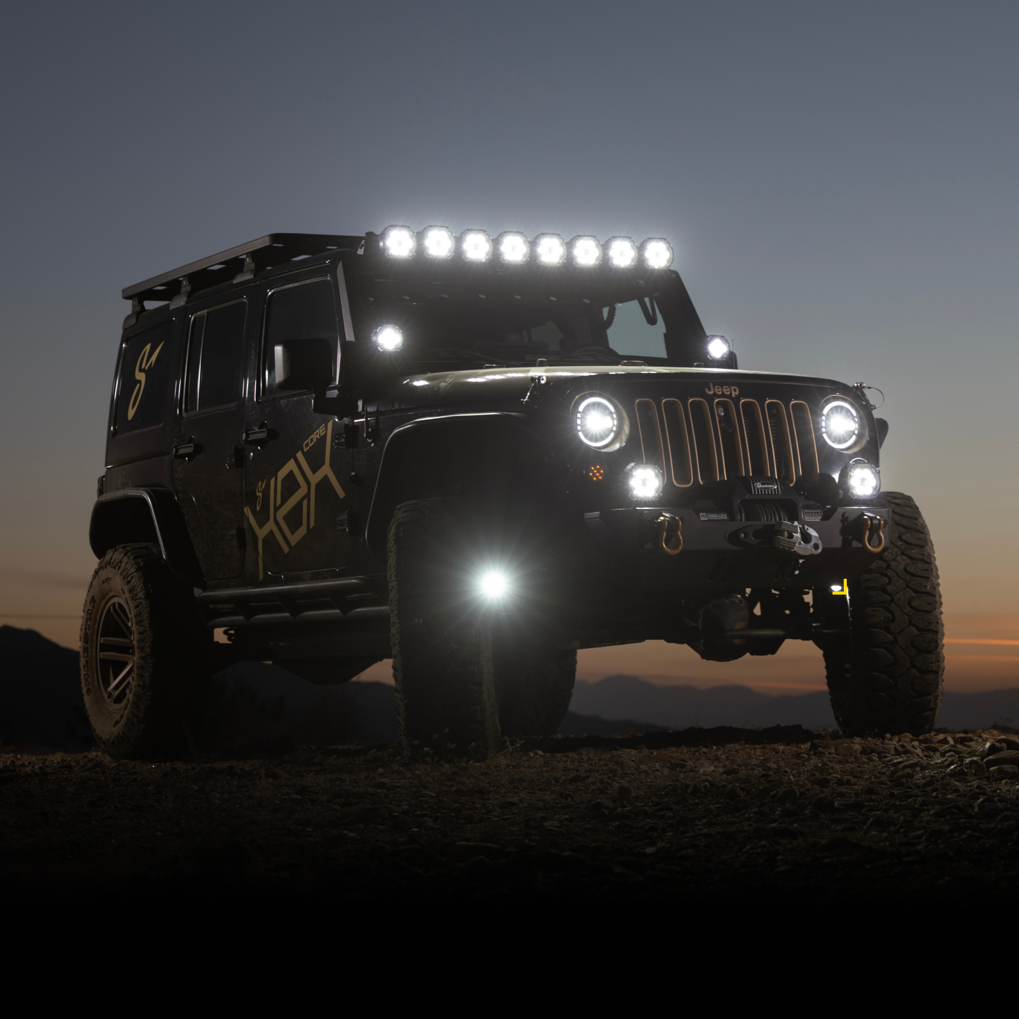 Black Jeep Wrangler with off-road lights and roof rack parked on a dirt trail at dusk