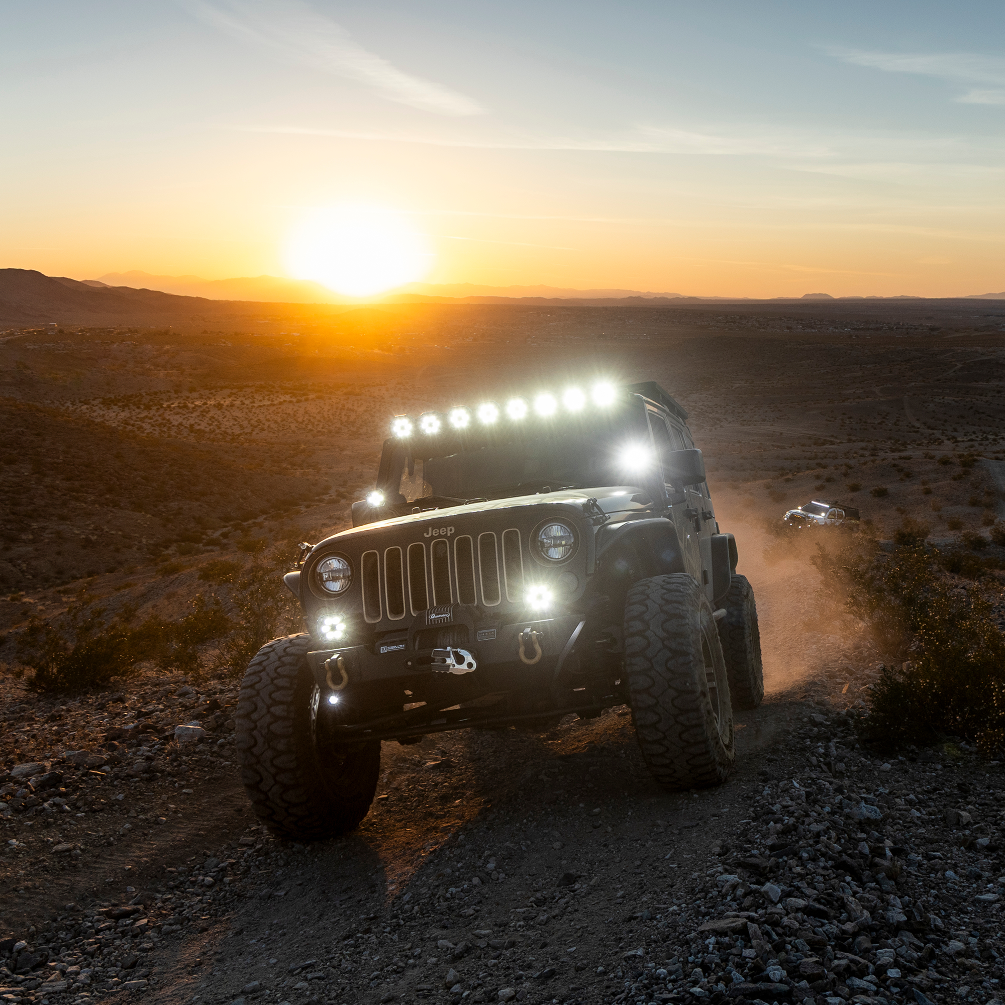 Jeep Wrangler driving on a rocky desert trail at sunset with LED light bar and auxiliary lights illuminated