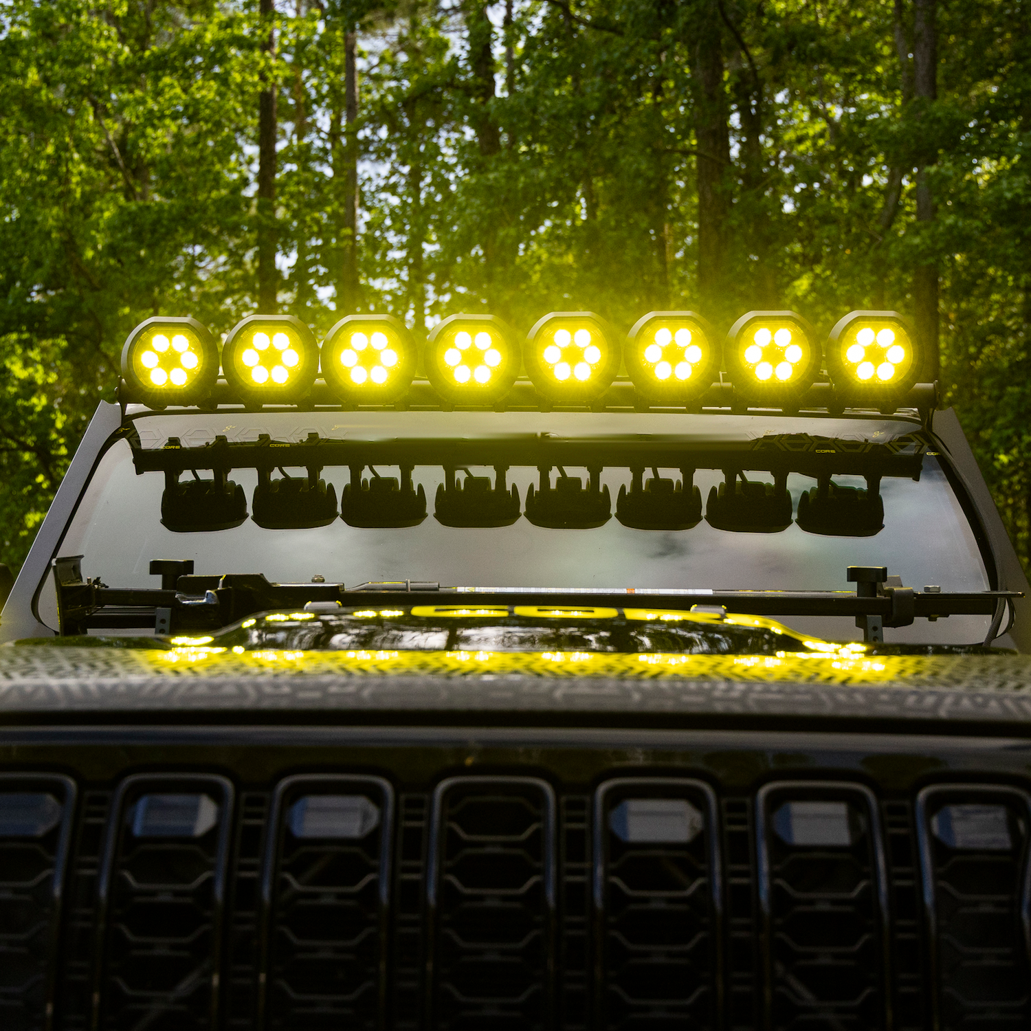 A row of bright yellow LED light bars mounted on the front windshield of an off-road vehicle, with a forest background.