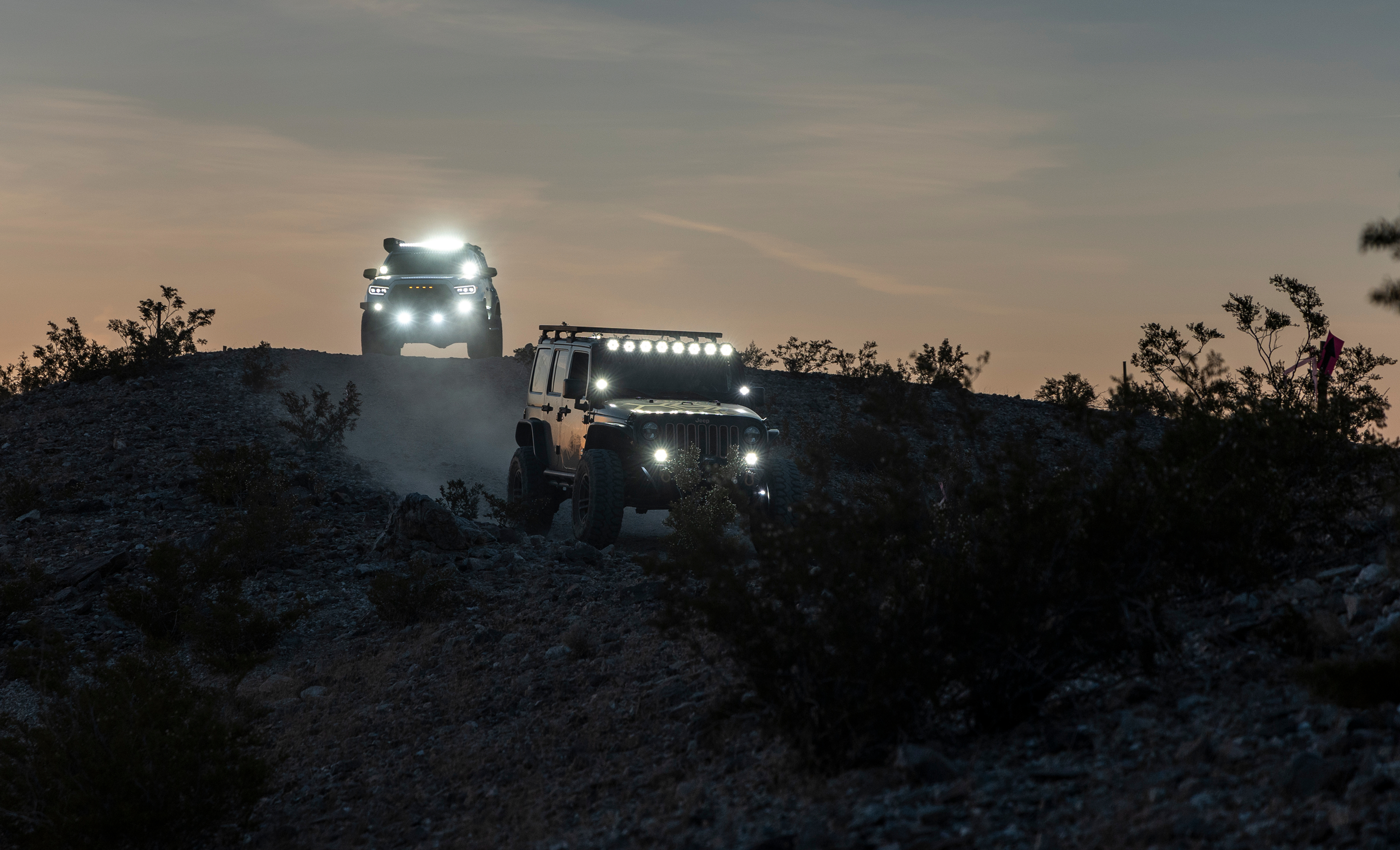 Two off road vehicles with bright LED lights driving on a desert ridge at dusk