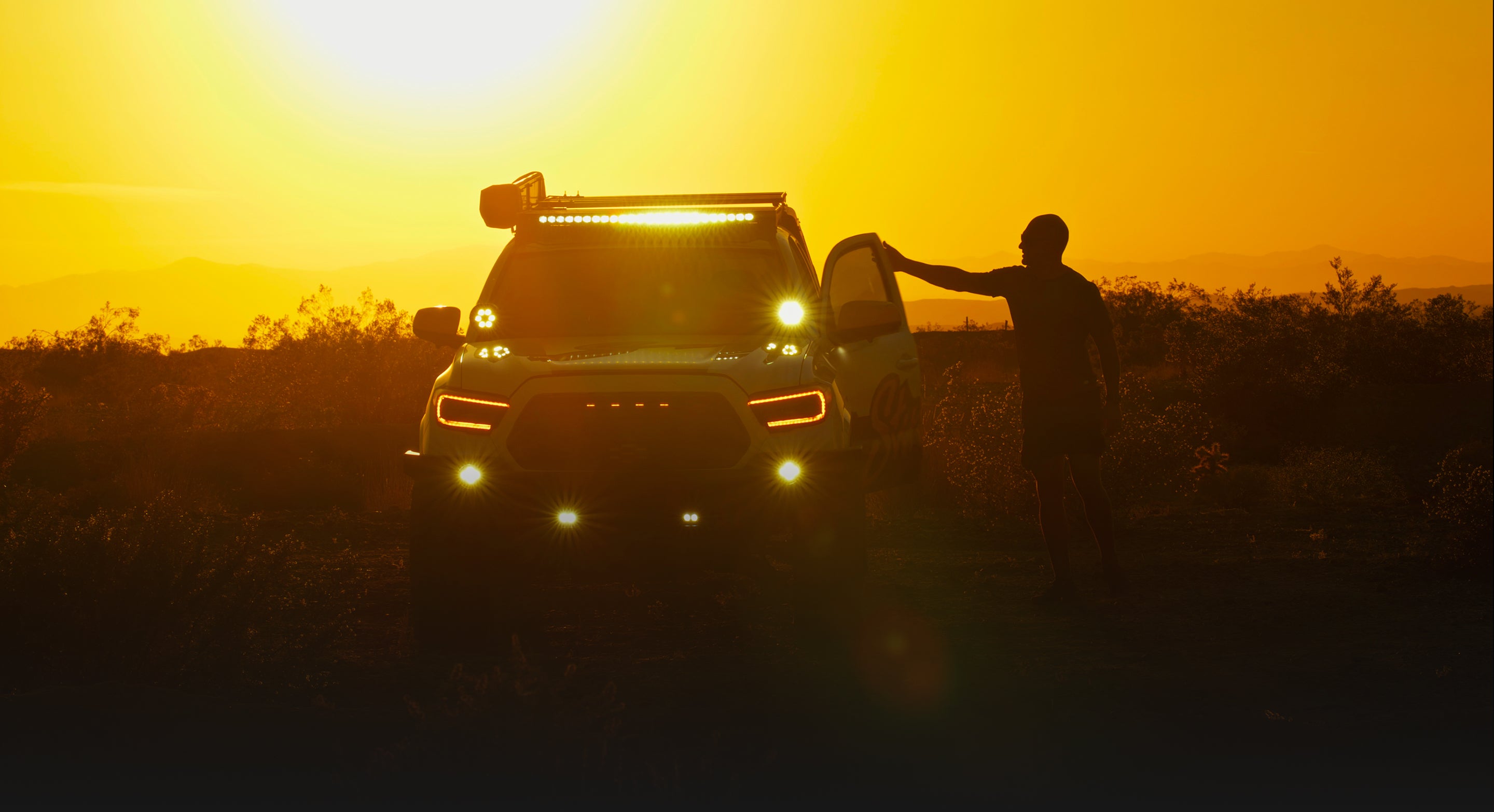 Person standing next to a vehicle with illuminated headlights in a field at sunset.