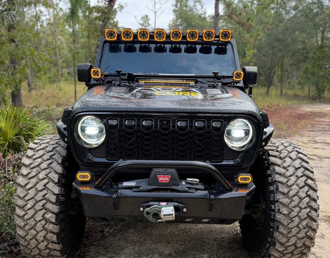 A black off-road Jeep with a custom black front bumper, winch, and a roof-mounted LED light bar with orange accents, parked on a dirt path with trees in the background.