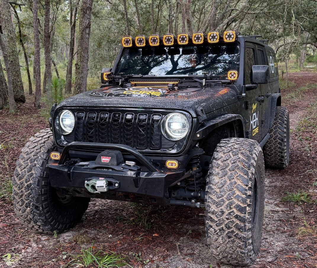 A black off-road Jeep with large knobby tires, a front bumper guard, and an auxiliary roof light bar, parked on a dirt trail in a wooded forest setting.