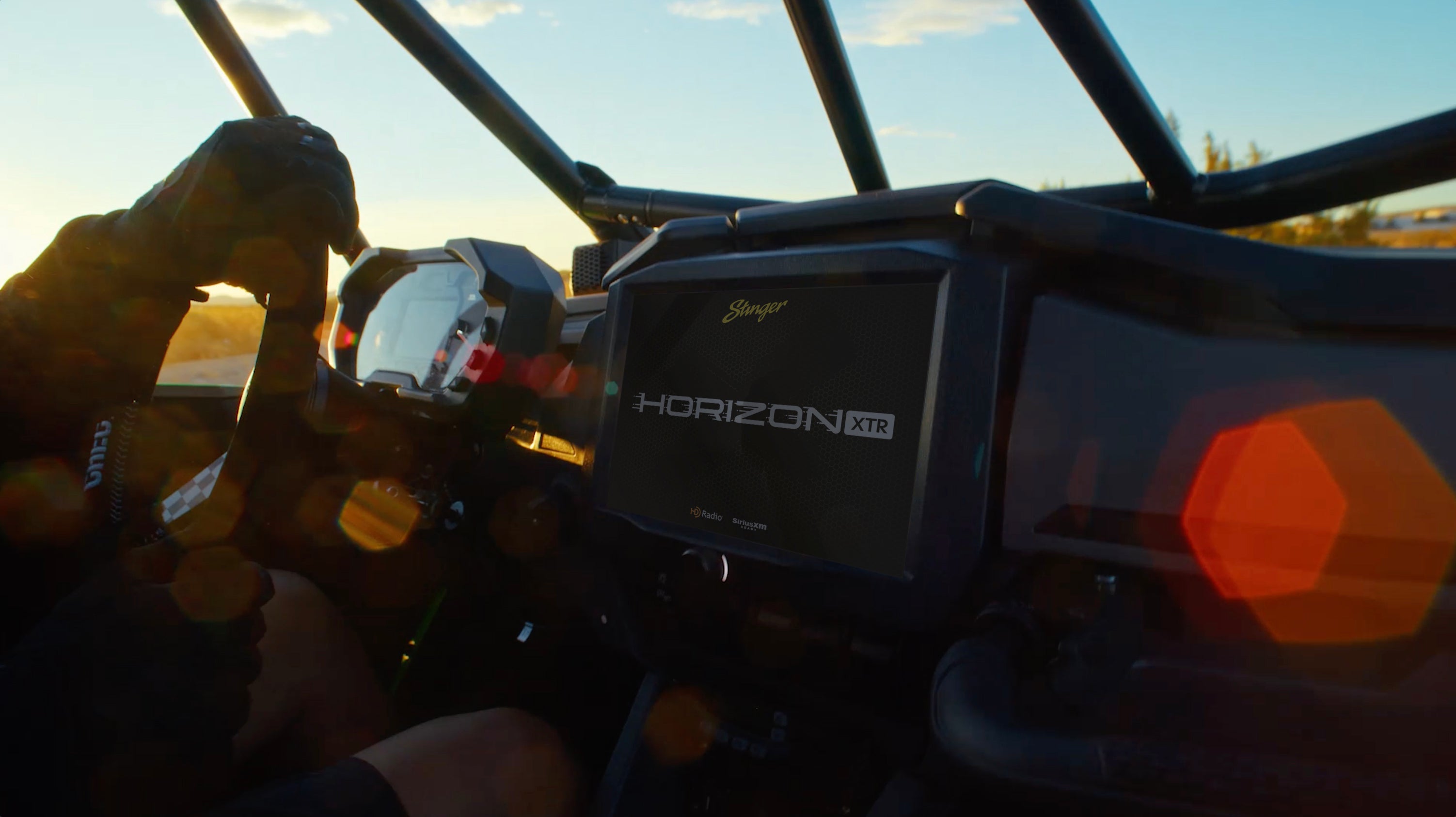 Stinger HORIZON XTR display installed in a UTV dash, with a gloved driver at the wheel under a desert sunset roll cage view