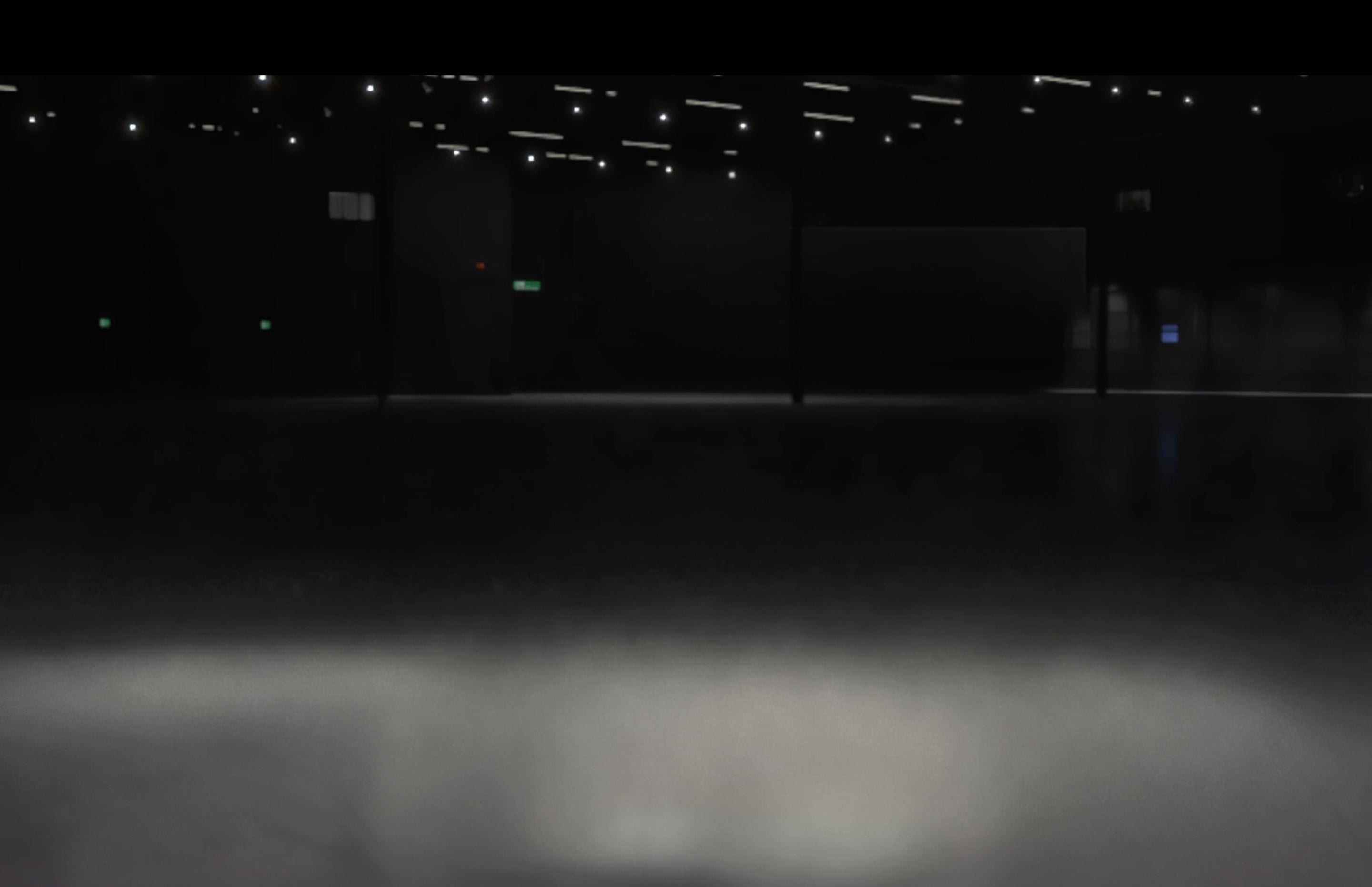 A dark, low-lit interior space with a smooth floor reflecting light from ceiling fixtures, and a bright horizontal band of illumination across the foreground.