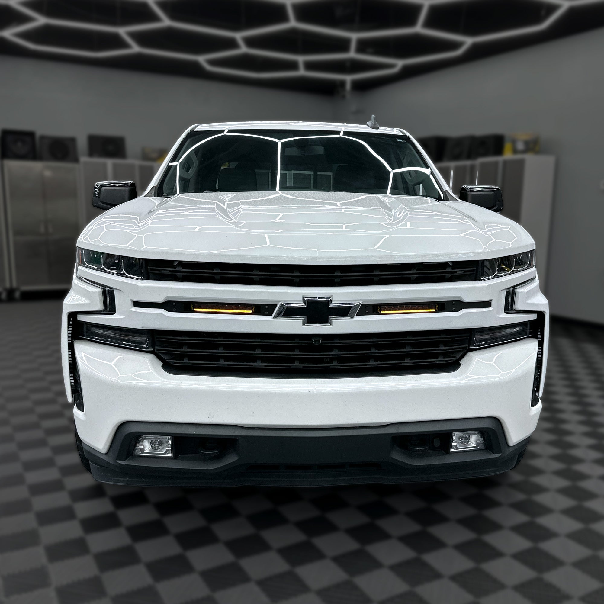 A white Chevrolet SUV photographed head-on in a clean indoor showroom or garage with a dark checkered floor and geometric ceiling lights.