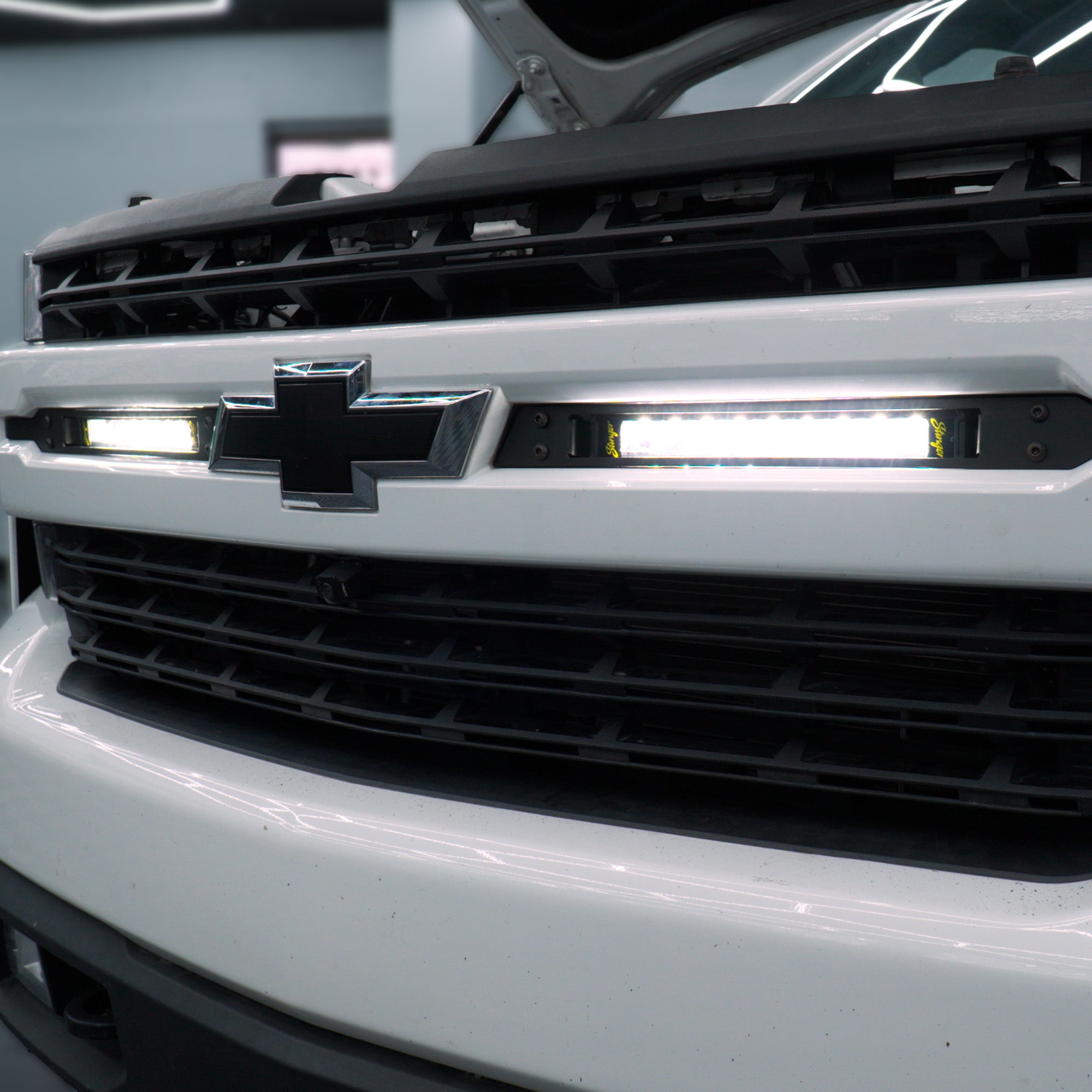 Close-up of a white Chevrolet truck front grille with integrated LED light bars illuminated, showing the Chevrolet bowtie logo and black grille details.