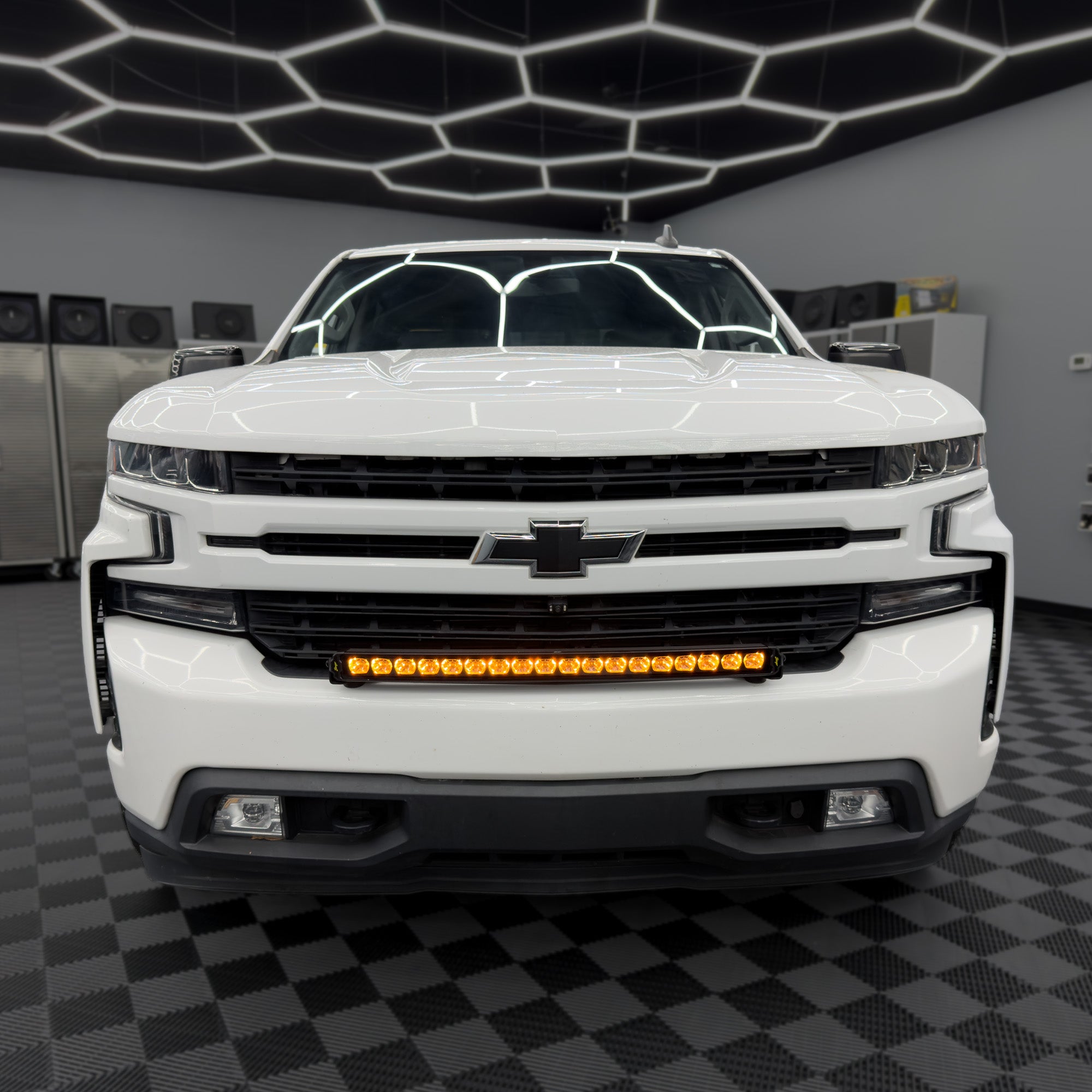 A white Chevrolet pickup truck shown from the front in a showroom, with an amber LED light bar mounted across the grille area.