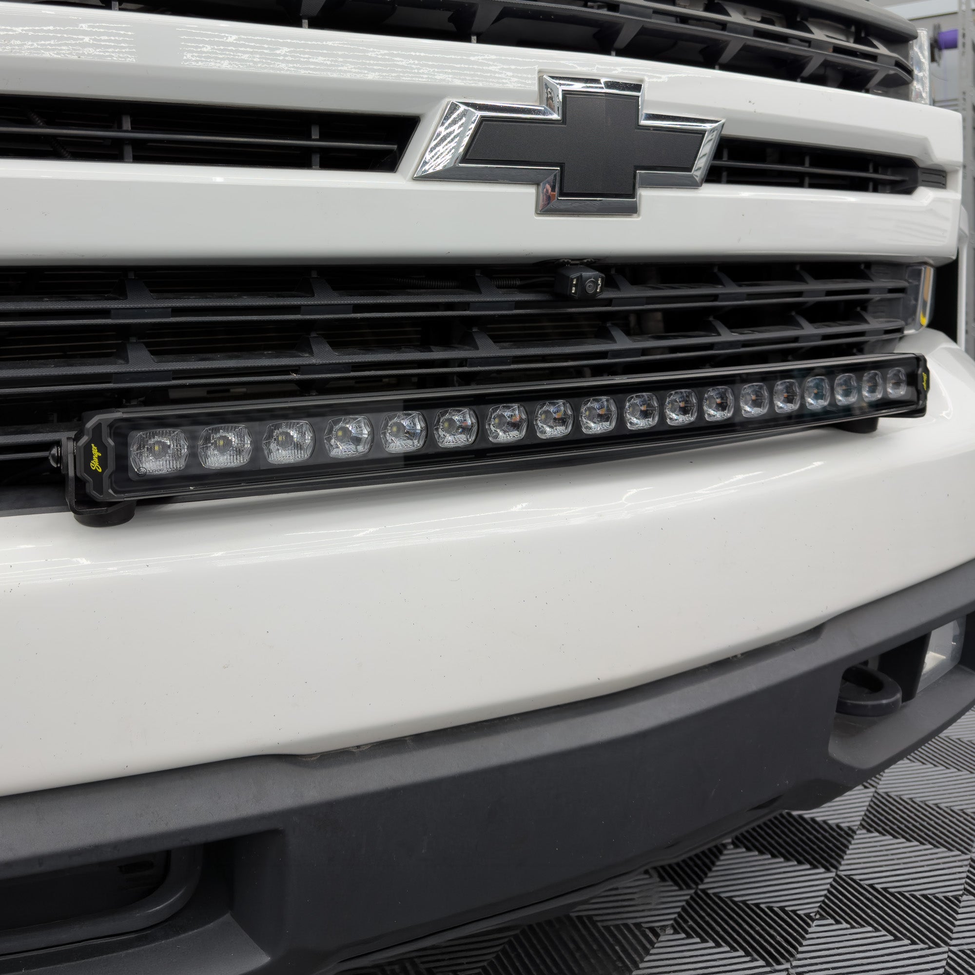 A black LED light bar mounted on the front bumper of a white Chevrolet truck, positioned below the chrome grille.