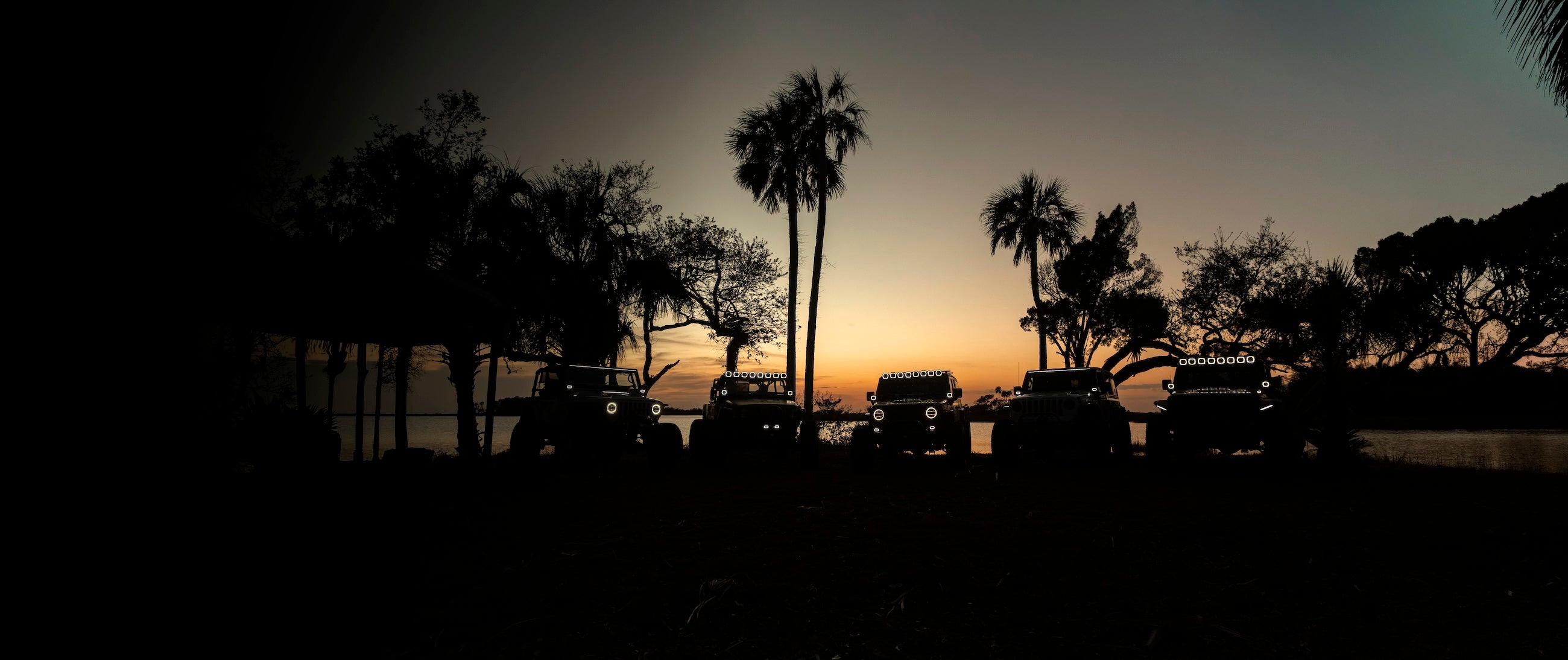 Five off road vehicles parked in silhouette against a sunset by a lake