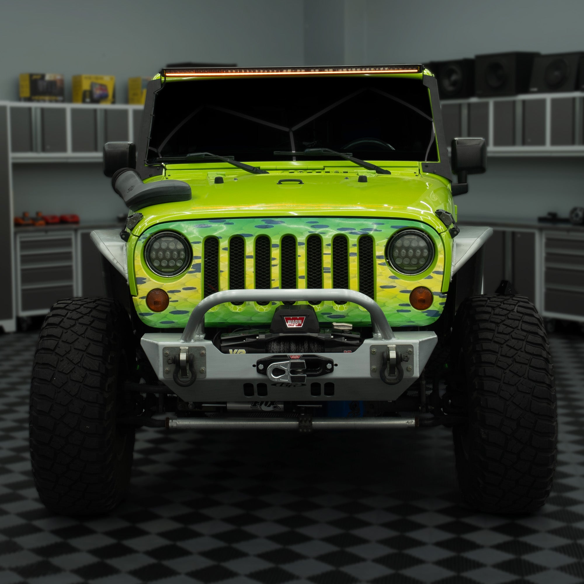 A customized lime green Jeep Wrangler with a camouflage-patterned front bumper and silver tubular front bar, parked in a garage with a black and white checkered floor.