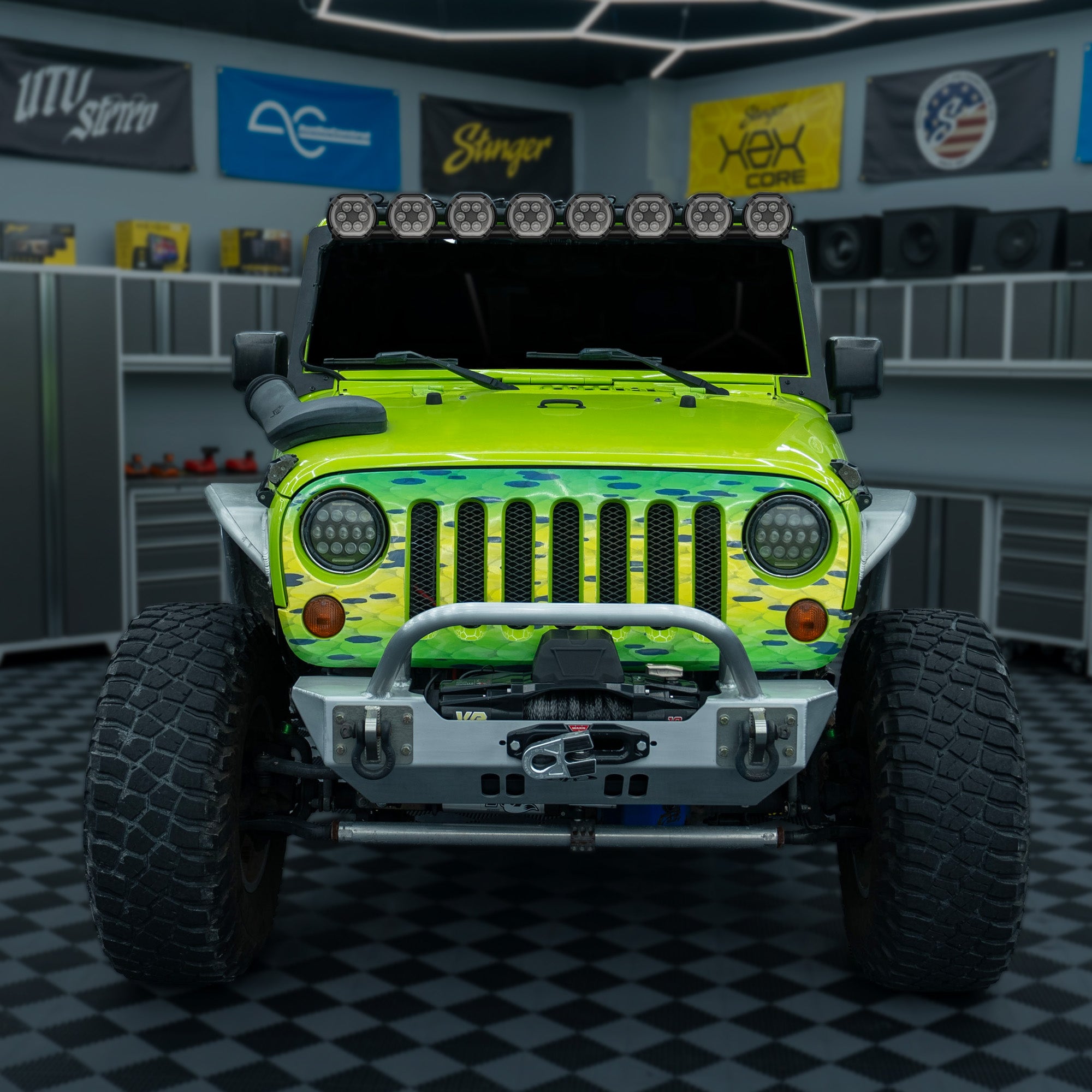 A customized lime green Jeep Wrangler with a camouflage pattern on the hood and fenders, parked inside a garage with a checkered floor and automotive decor.