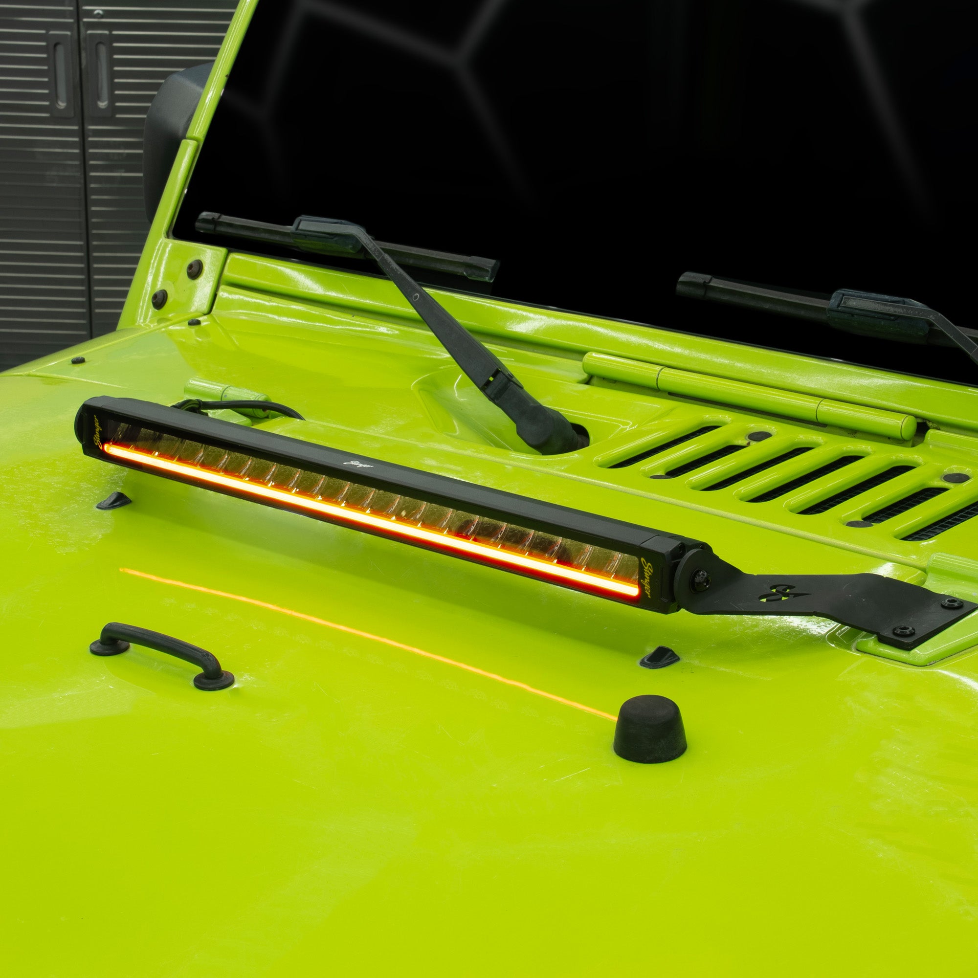 A long rectangular LED light bar with a black housing and mounting brackets, mounted on the hood of a bright lime green vehicle. The light bar is illuminated, casting a warm orange glow.