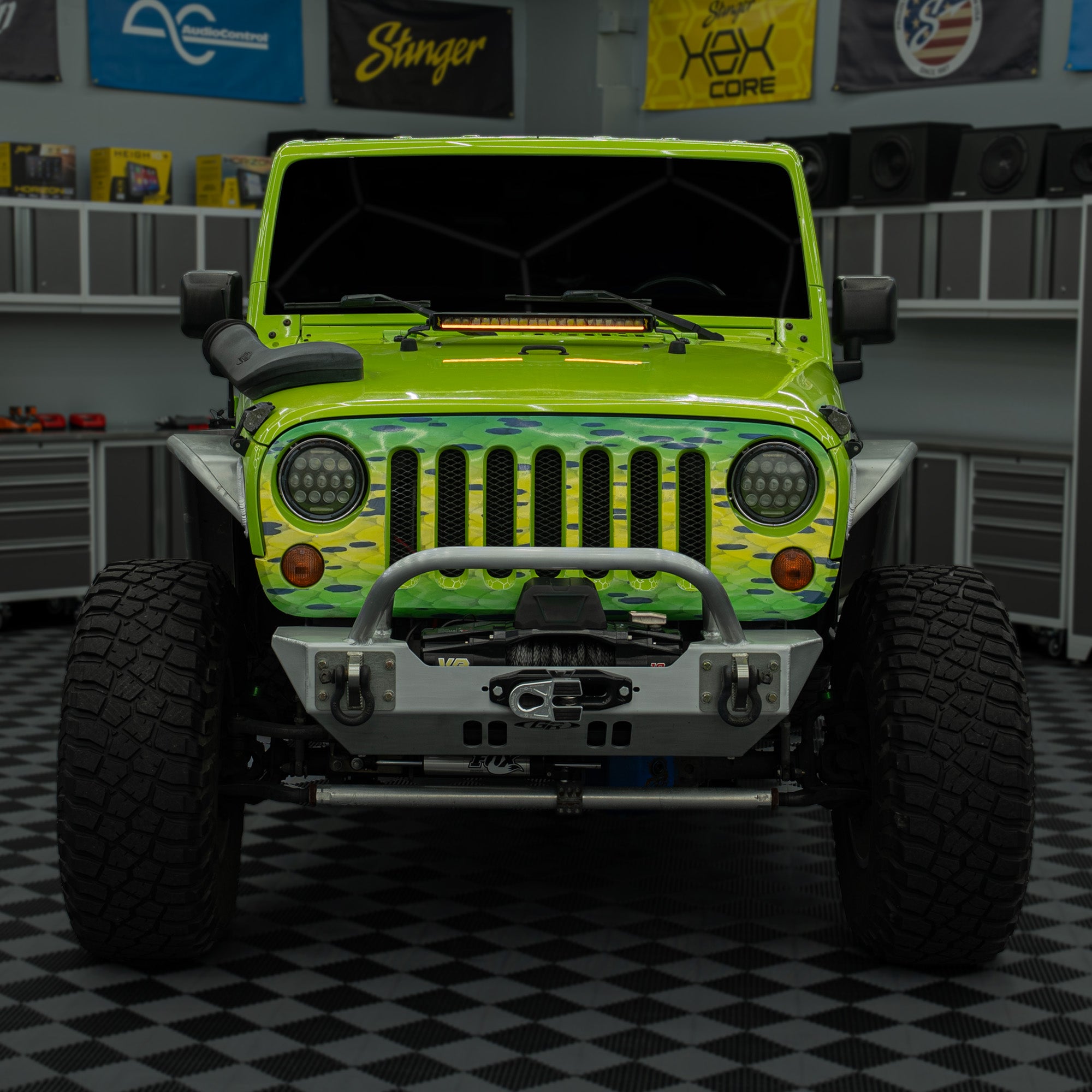 A customized lime green Jeep Wrangler with a camouflage wrap on the hood and fenders, parked in a garage setting with a checkered floor.