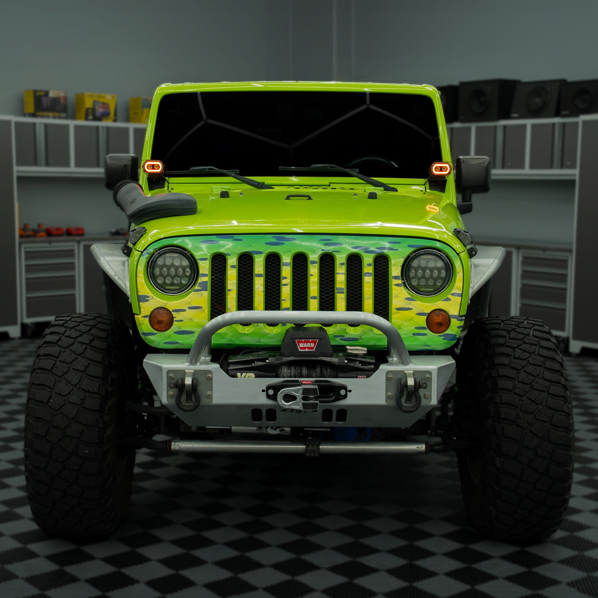 A lime green Jeep Wrangler with a camouflage front grille decal, a silver front bumper, and off-road tires, parked inside a garage with a black and white checkered floor.