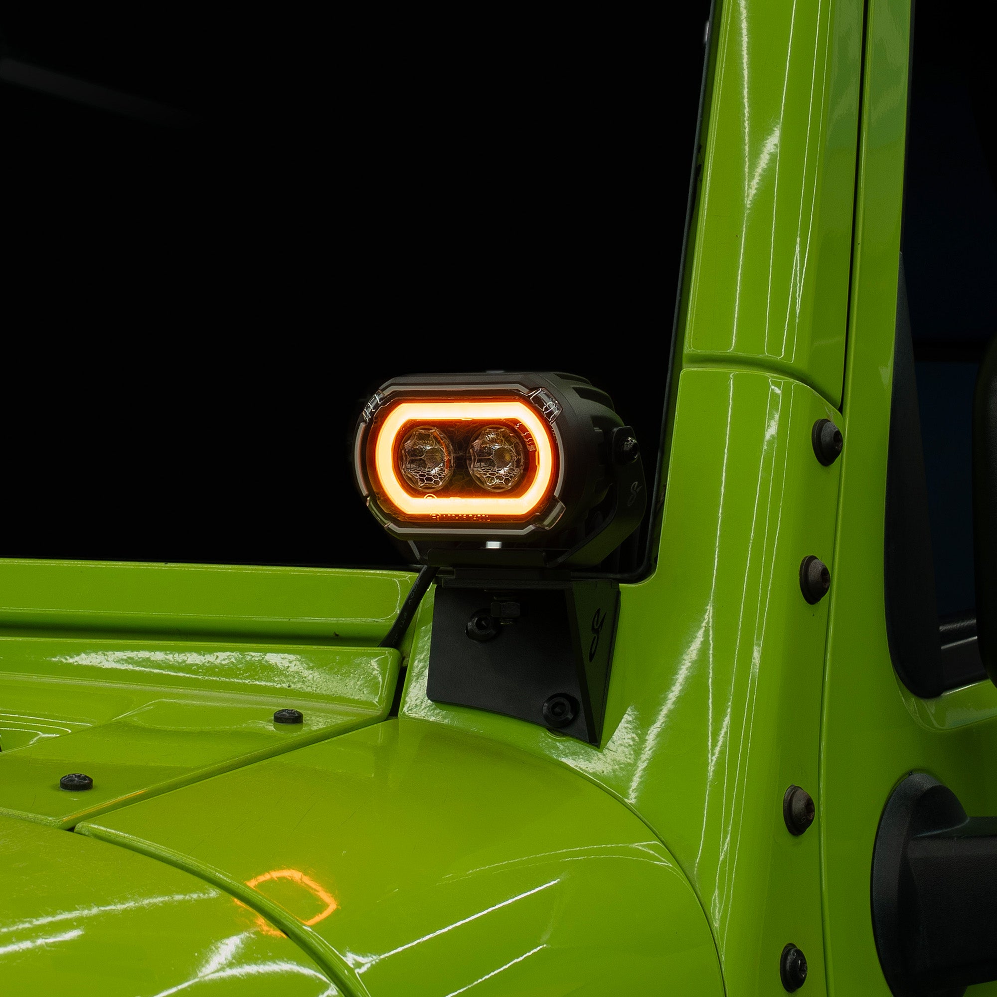 A close-up of a black rectangular LED light with a glowing amber ring, mounted on the side of a bright lime green vehicle against a dark background.