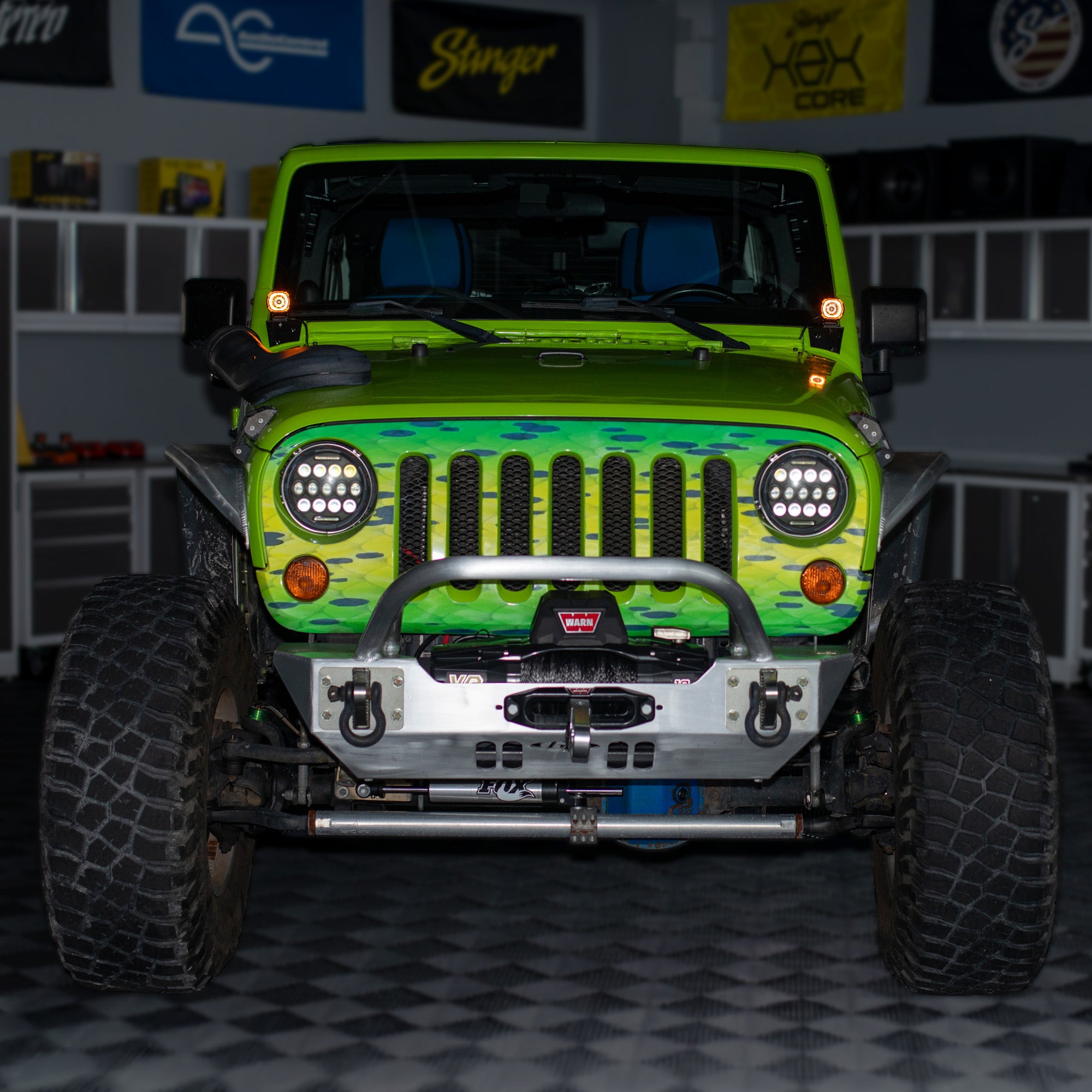 A customized lime green Jeep Wrangler with a green camouflage front grille wrap, a heavy-duty silver front bumper, and large off-road tires, parked in a garage.