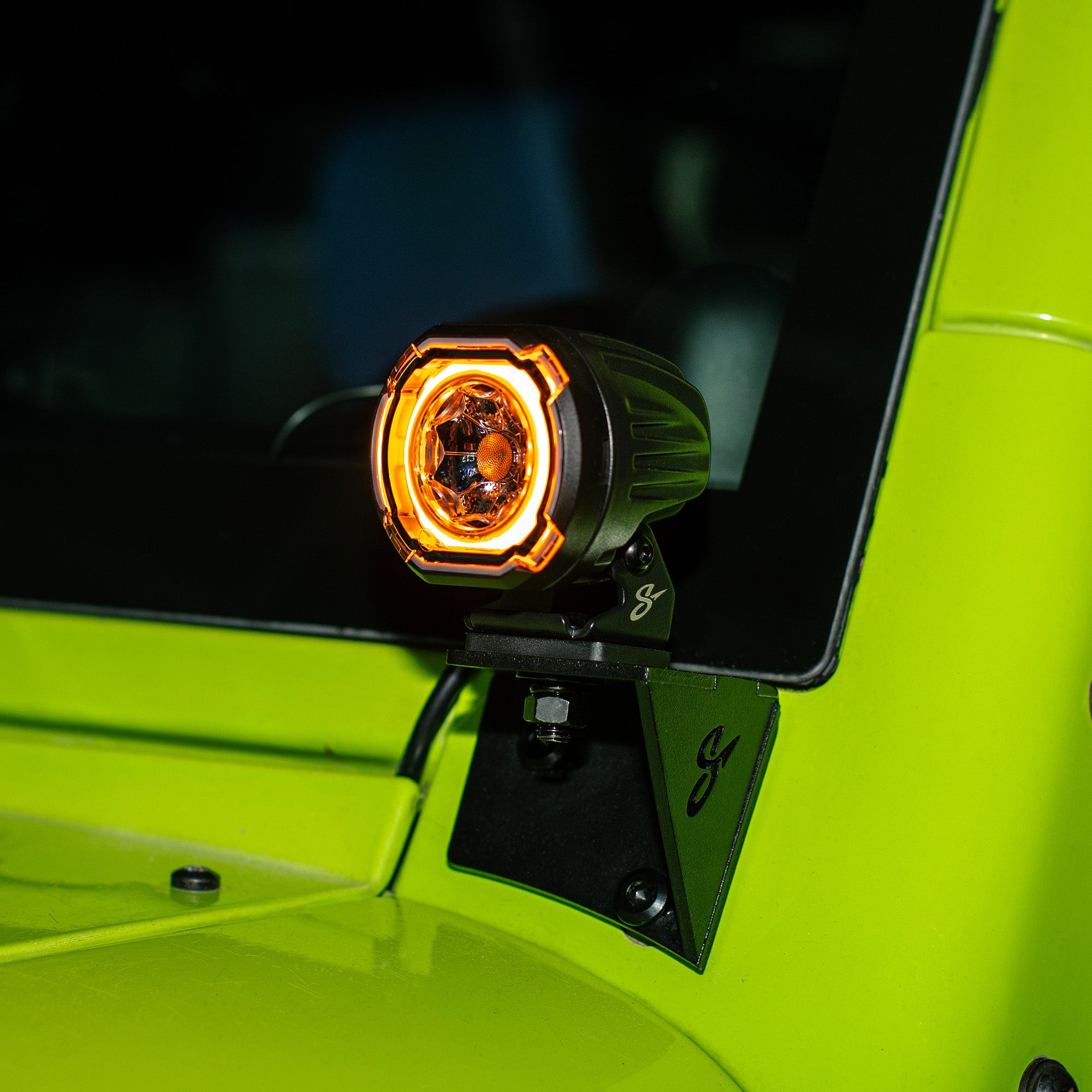 A black off-road LED light with an illuminated orange ring, mounted on a black metal bracket attached to the bright lime green body of a vehicle.