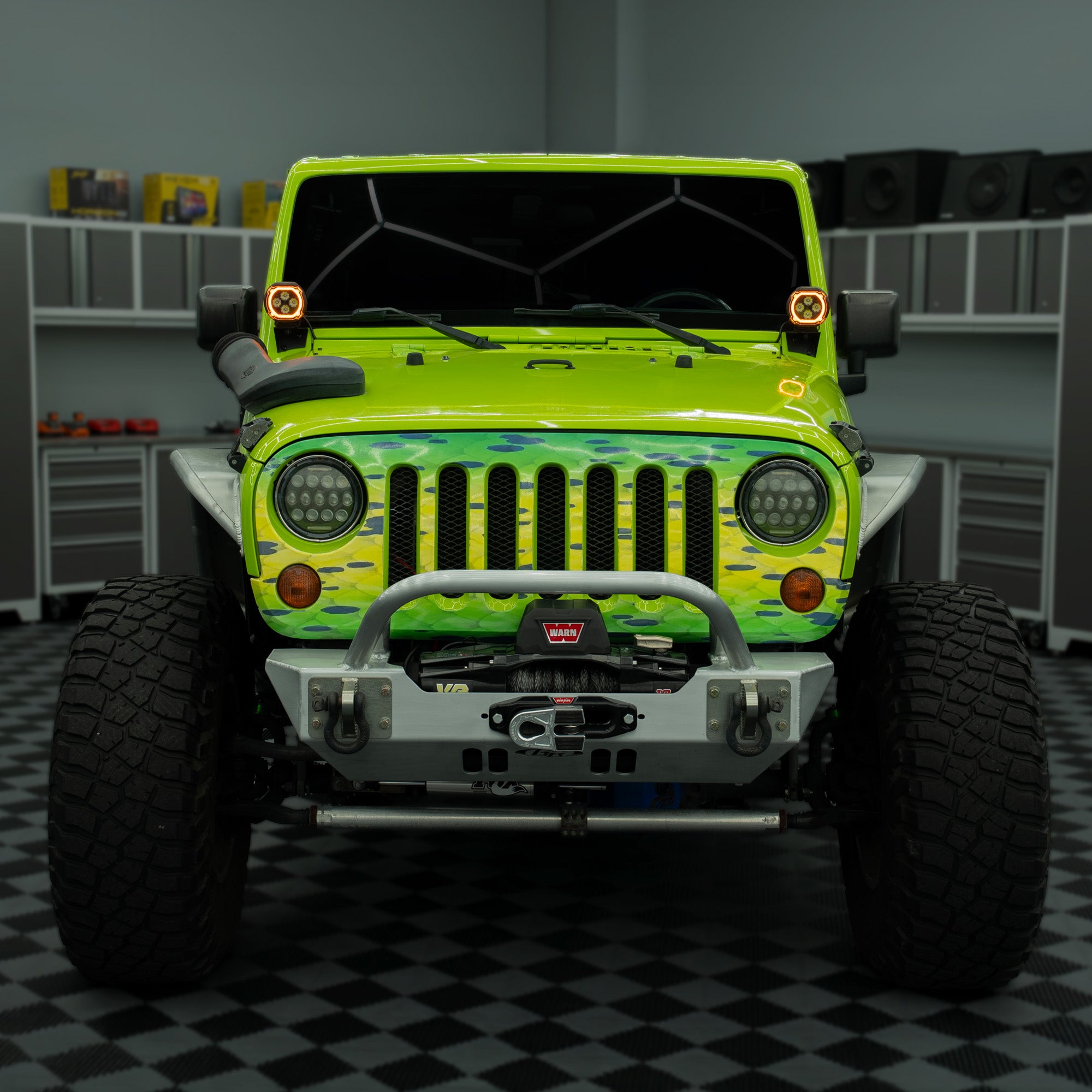 A customized lime green Jeep Wrangler with a green camouflage wrap on the hood and fenders, a silver front bumper guard, and large off-road tires, parked in a garage setting.