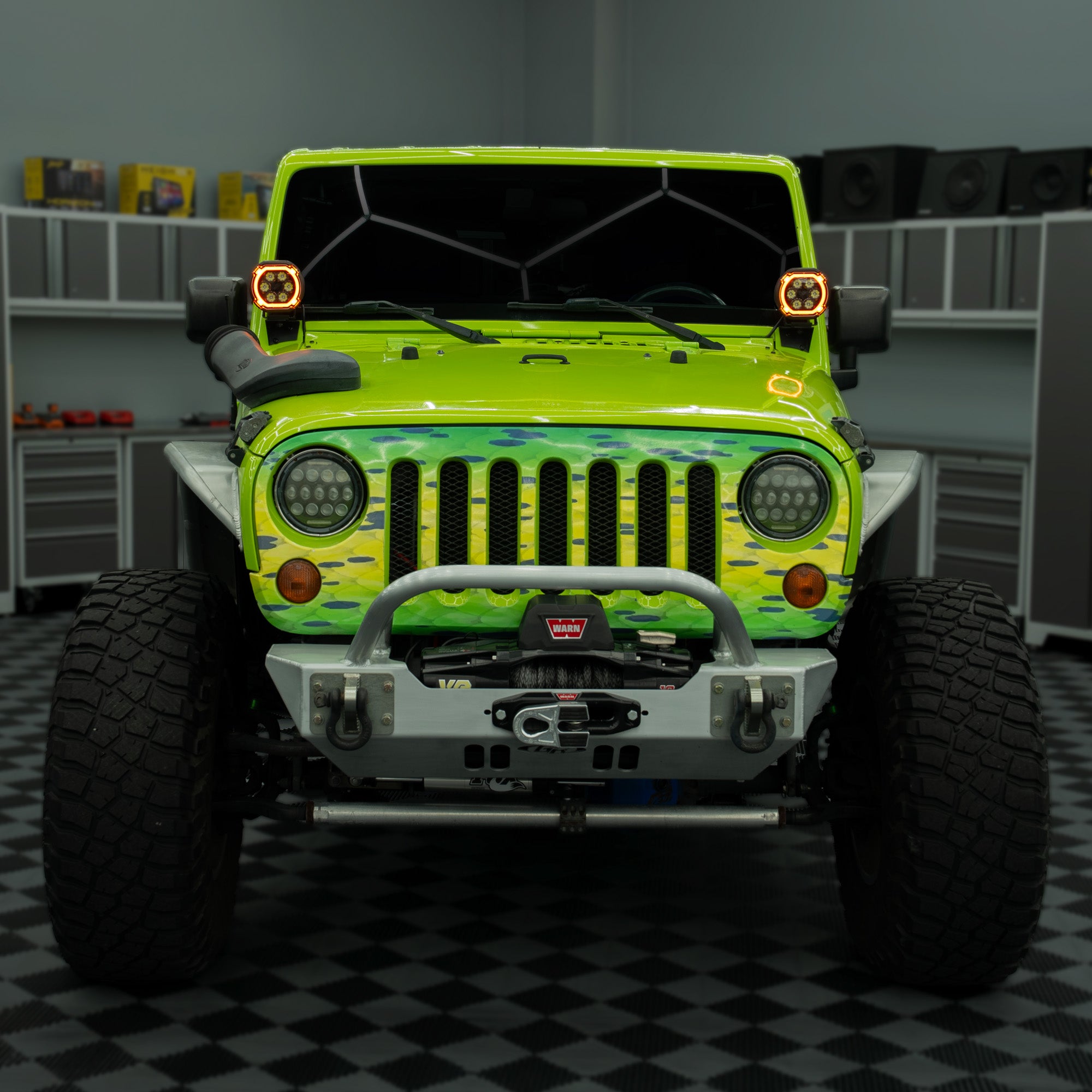 A bright lime green Jeep Wrangler with a green camouflage front grille wrap, a silver front bumper, and large off-road tires, shown from the front in a garage setting.