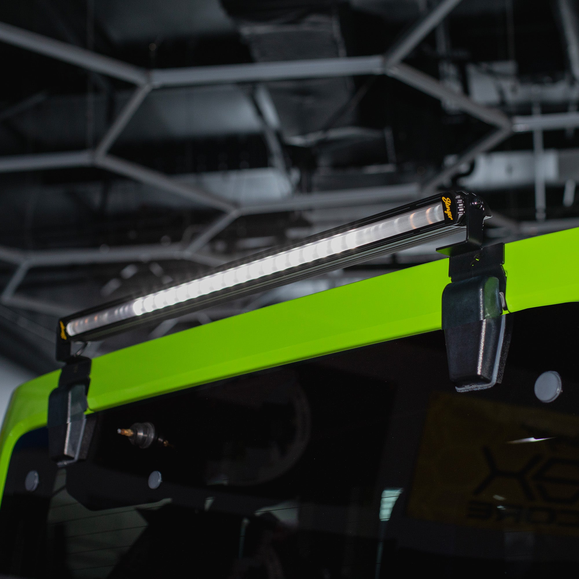 A bright green roof-mounted LED light bar clipped onto the roof rack of a vehicle, shown in a dark indoor setting with a blurred geometric ceiling structure in the background.