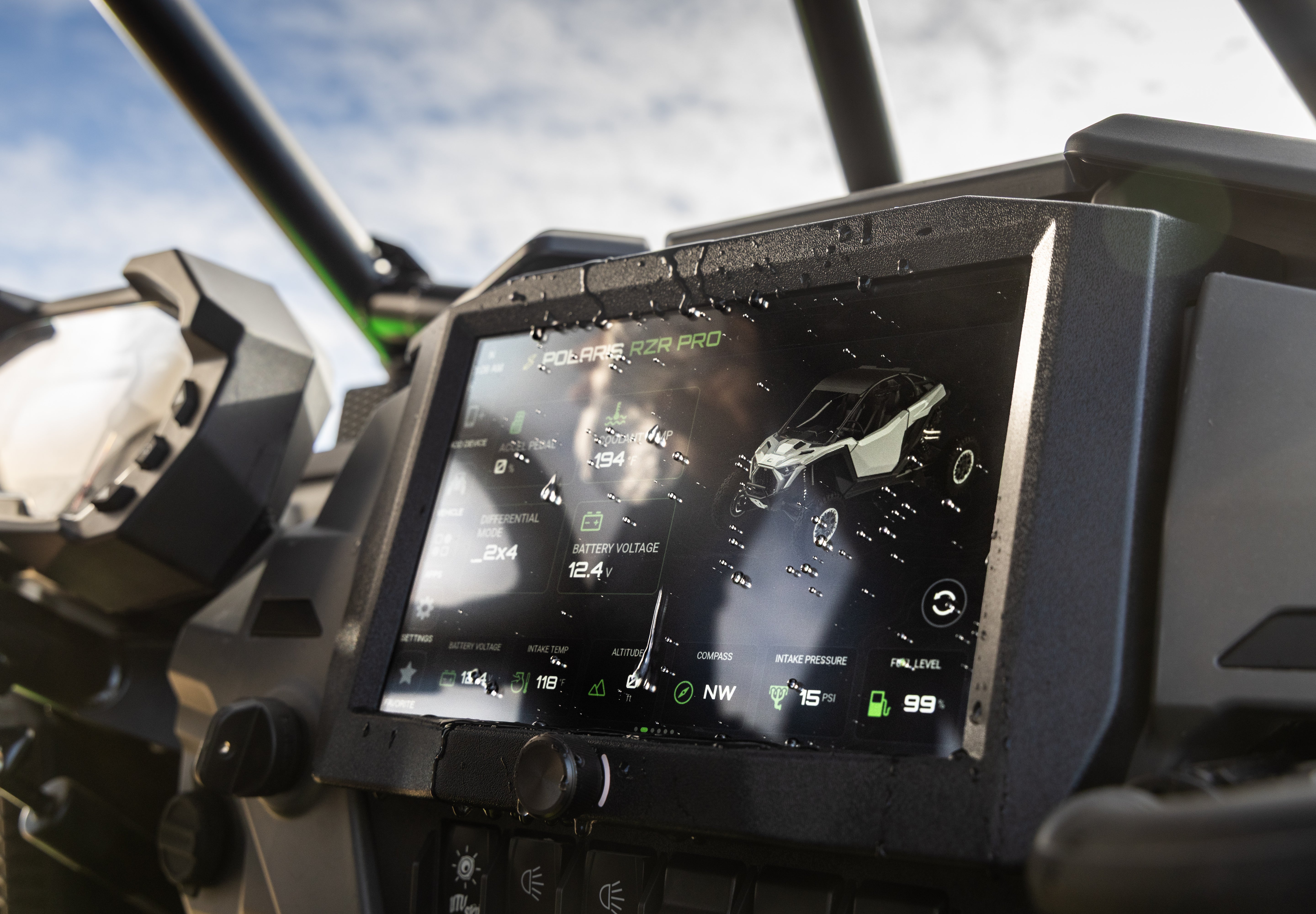 A close-up of a rugged touchscreen display mounted on a vehicle dashboard, showing a digital instrument interface. The screen, set against a dark console with outdoor sky visible through the windshield.