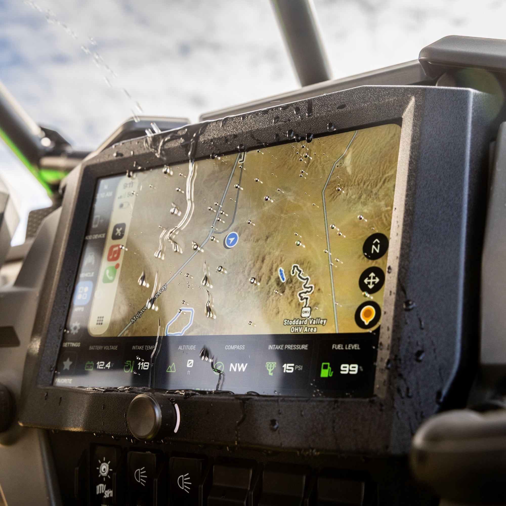 A rugged touchscreen display mounted on a vehicle dashboard, showing a map and vehicle status icons. The screen is covered with water droplets, and the surrounding controls and roll bar are visible in an outdoor setting.