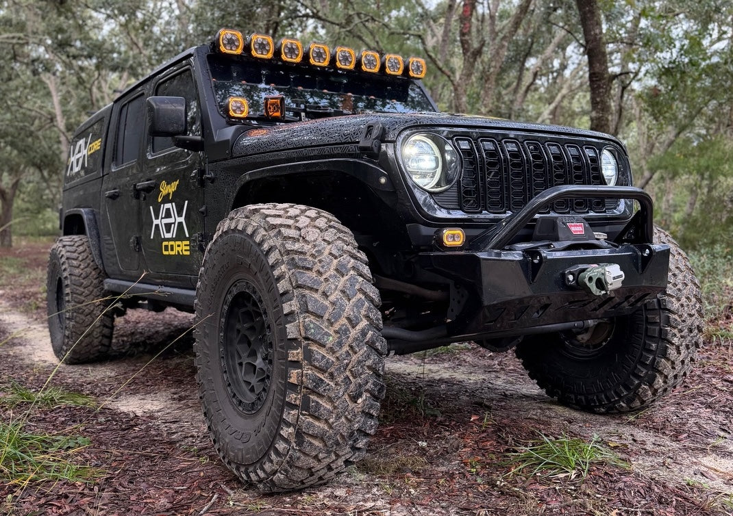 A black lifted off-road vehicle with large knobby tires, a front bull bar, roof-mounted LED light bar, and custom graphics, photographed on a dirt trail surrounded by trees.